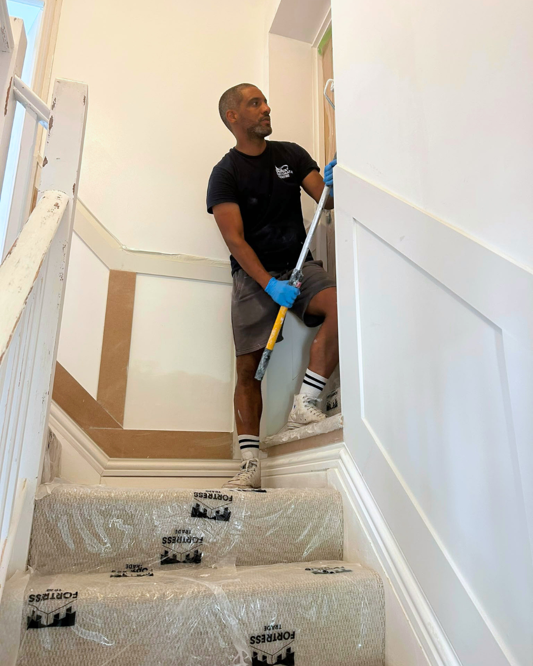 Painter applying fresh paint to a hallway and landing staircase in Renhold, with protective sheeting on the stairs.