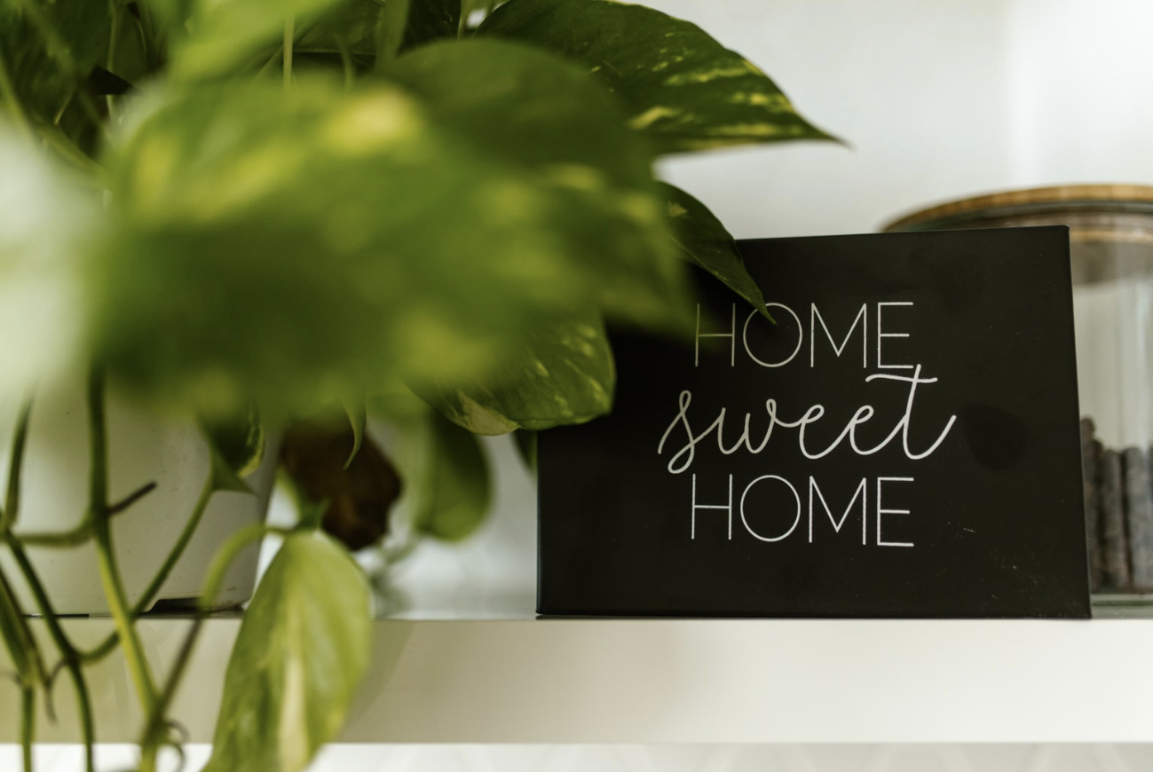 A 'Home Sweet Home' sign on a shelf beside a houseplant.